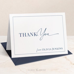 May include: A white thank you card with a navy blue border and text. The card says "Thank You" in a script font with a navy blue line underneath. Below the text, it says "from Olivia Jenkins" in a smaller font.
