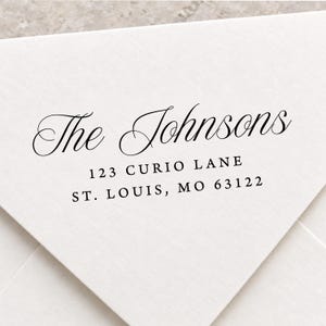 May include: A white envelope featuring black printed text that reads The Johnsons, 123 Curio Lane, St. Louis, MO 63122. The name is written in a flowing script font, while the address is displayed in a clean, simple serif typeface.