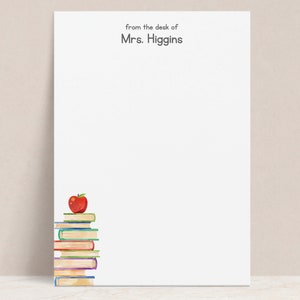 May include: White stationery with the text "from the desk of Mrs. Higgins" at the top. A stack of colorful books with a red apple on top is in the lower left corner. The stationery is blank and ready for writing.