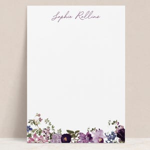 May include: White stationery with a floral border featuring purple, lavender, and blue flowers. The name "Sophie Rollins" is written in a cursive font at the top of the page. The stationery is blank, offering space for personalized messages.