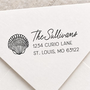 May include: A black ink rubber stamp impression on a white envelope featuring a detailed line drawing of a seashell on the left. The text reads The Sullivans, 1234 CURIO LANE, ST. LOUIS, MO 63122 in a combination of script and sans-serif fonts.