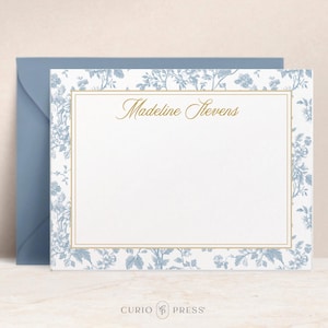 May include: Stationery set featuring a personalized note card and envelope. The note card has a white background with a gold border and the name "Madeline Stevens" in gold script. The floral design is blue and white.