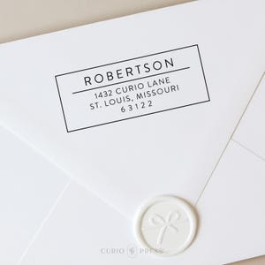 May include: A white envelope featuring a black rectangular stamp with the text ROBERTSON, 1432 CURIO LANE, ST. LOUIS, MISSOURI 63122. The envelope is sealed with a circular white wax seal embossed with a simple line drawing of a bow.