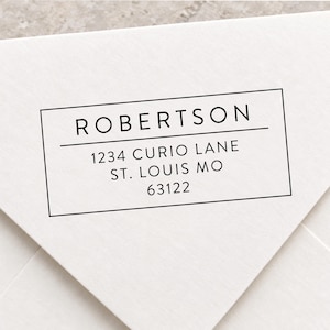 May include: A rectangular black ink stamp impression on a white envelope. The text reads ROBERTSON, followed by a horizontal line, then 1234 CURIO LANE, ST. LOUIS MO, and 63122. The design is enclosed within a thin black rectangular border.