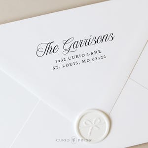 May include: White envelope with the address "The Garrisons" and the address "1432 Curio Lane St. Louis, MO 63122" printed in black. A white wax seal with a bow design is on the bottom right corner. The Curio Press logo is at the bottom.