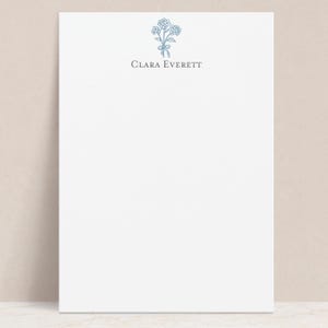 May include: White stationery with a blue floral illustration and the name "Clara Everett" printed in a serif font. The floral design is a cluster of flowers tied with a ribbon. The paper is blank, with a simple, elegant design.