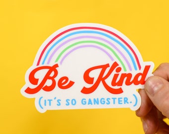 May include: A sticker with a rainbow design and the words "Be Kind" in red, with the phrase "(it's so gangster.)" in blue. The sticker is held against a yellow background.