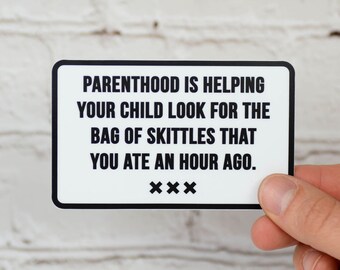 May include: A white rectangular sticker with rounded corners, featuring black text that reads: "PARENTHOOD IS HELPING YOUR CHILD LOOK FOR THE BAG OF SKITTLES THAT YOU ATE AN HOUR AGO." The sticker is held by a hand.