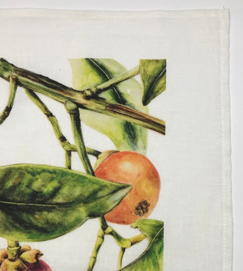 May include: A close-up of a white linen napkin with a printed design of a branch of mangosteen fruit with green leaves. The fruit is orange and red with a dark brown center.