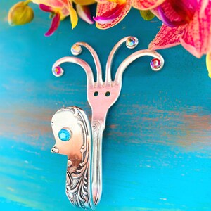 May include: A silver fork has been transformed into a quirky wall hook, featuring blue rhinestones. The hook is shaped like a bird with a long, curved neck and an ornate design. The hook is mounted on a turquoise blue wall.