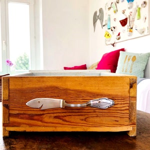 Op de afbeelding: Een houten kist met een zilveren visvormige handgreep. De kist is gemaakt van lichtbruin hout en staat op een donkerbruine tafel. De handgreep is een hergebruikte lepel met een vissenkopontwerp. De kist lijkt een decoratief opbergitem te zijn.