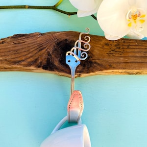 May include: A silver spoon with a decorative swirl design is attached to a piece of driftwood. The driftwood is mounted on a light blue wall. A white orchid is visible in the background.