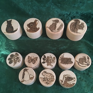 May include: Twelve wooden round ornaments with various animal and nature designs, including a cat, rabbit, mouse, squirrel, berries, butterfly, flowers, hedgehog, frog, and badger.