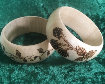 May include: Two wooden bangles with a floral design burned into the wood. The design features leaves and berries.