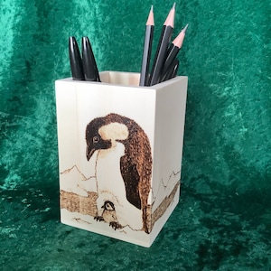 May include: A white wooden pencil holder with a woodburned image of two penguins on a snowy landscape. The holder has four pencils inside.