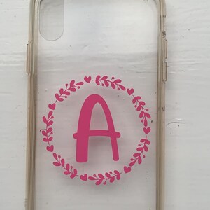 Op de afbeelding: Een transparante telefoonhoes met een roze bloemenkrans en de letter "A" in het midden.