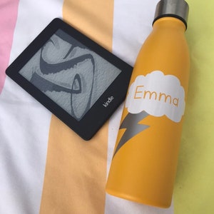 Puede incluir: Una botella de agua amarilla con una nube blanca y un diseño de rayo. La botella tiene el nombre "Emma" escrito en ella. También se ve un lector electrónico Kindle en blanco y negro en la imagen.