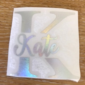 Op de afbeelding: Een witte sticker met een holografische afwerking met de letter "K" en de naam "Kate" in een cursief schrift.