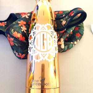 May include: A rose gold reusable water bottle with a white monogram and a white mandala design. The bottle has the text "one green bottle" printed on the bottom.