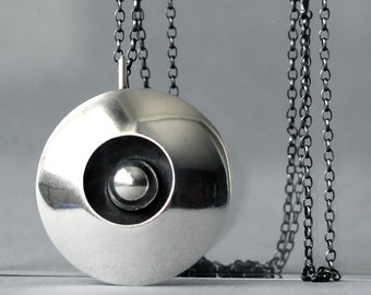 May include: A silver pendant necklace with a round, concave disc featuring a smaller, raised circle in the center. The necklace is on a black chain.
