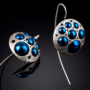 May include: Silver-toned earrings with a textured, circular design. Each earring features multiple, glossy, deep blue orbs of varying sizes, set within the silver framework. The earrings have a long, curved hook.