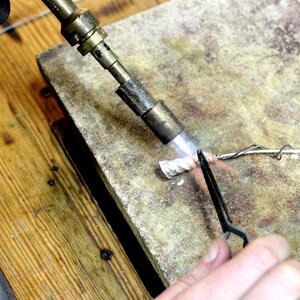 K&ouml;nnte beinhalten: Eine Person verwendet einen Brenner, um ein mit einer Pinzette auf einer grauen Oberfl&auml;che gehaltenes St&uuml;ck Metallschmuck zu erhitzen.