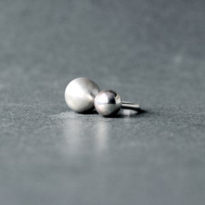 Può includere: Un anello d'argento con due sfere rotonde e lisce. La sfera più grande è a sinistra e la più piccola a destra.