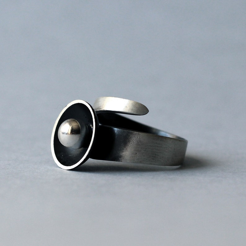 May include: A silver ring featuring a black and silver disc with a silver ball at its centre.