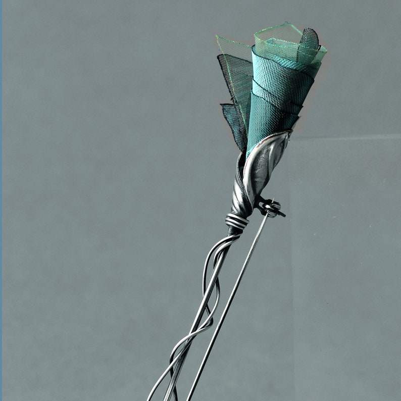 K&ouml;nnte beinhalten: Eine silberne Brosche in Form einer Rose mit einer blaugr&uuml;nen Stoffblume. Die Brosche ist an einer silbernen Nadel befestigt.