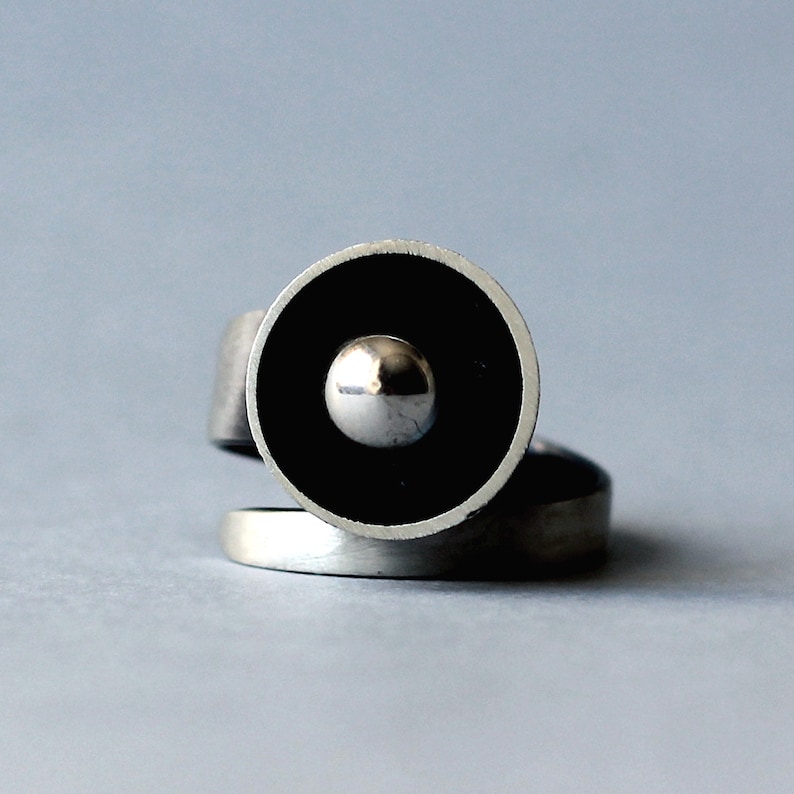 May include: A silver ring featuring a black circular design with a silver sphere at its centre.