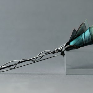 K&ouml;nnte beinhalten: Eine silberne Brosche mit einem filigranen, gedrehten Drahtdesign, das einem Stiel &auml;hnelt. Der Stiel ist mit einem B&uuml;schel aus t&uuml;rkisfarbenem und schwarzem Stoff bedeckt, der einer Blume &auml;hnelt.