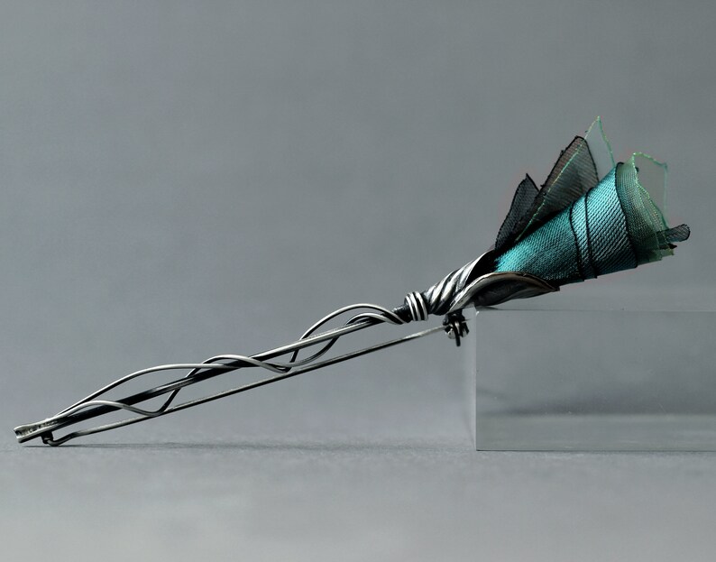 K&ouml;nnte beinhalten: Eine silberne Brosche mit einem filigranen, gedrehten Drahtdesign, das einem Stiel &auml;hnelt. Der Stiel ist mit einem B&uuml;schel aus t&uuml;rkisfarbenem und schwarzem Stoff bedeckt, der einer Blume &auml;hnelt.