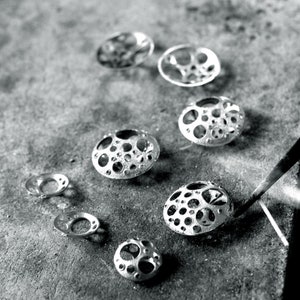 May include: A collection of silver metal jewelry pieces with a circular design featuring a pattern of small holes. The pieces are arranged in a row on a gray surface.