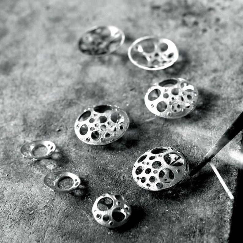 May include: A collection of silver metal jewelry pieces with a circular design featuring a pattern of small holes. The pieces are arranged in a row on a gray surface.