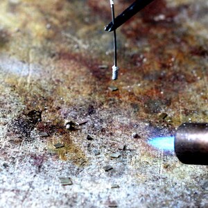 May include: A close-up of a metal object being heated with a blowtorch. The object is held in place by a pair of tweezers. There are small pieces of metal scattered on the surface.