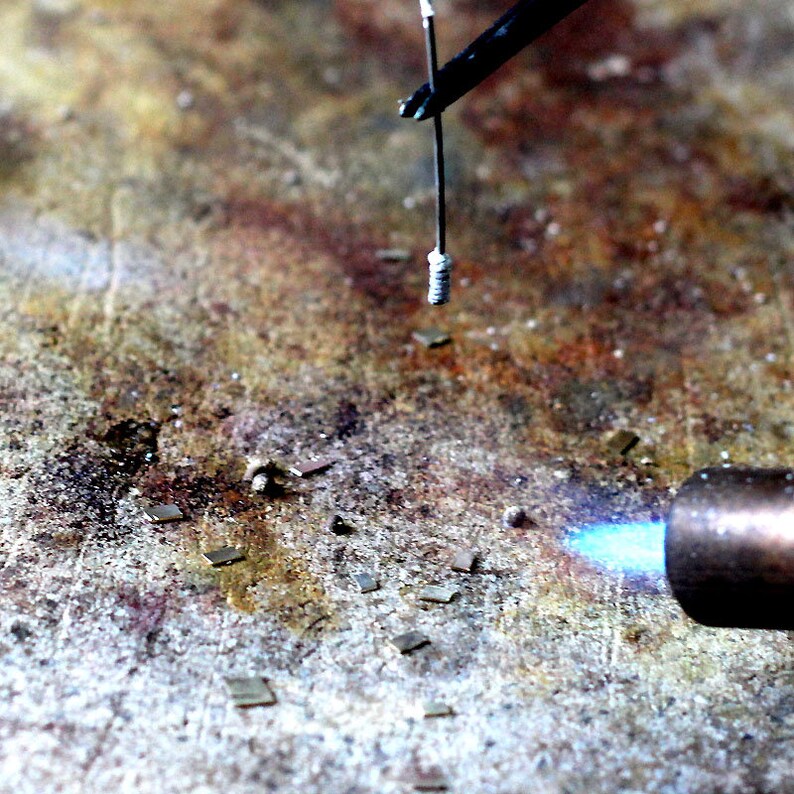 May include: A close-up of a metal object being heated with a blowtorch. The object is held in place by a pair of tweezers. There are small pieces of metal scattered on the surface.