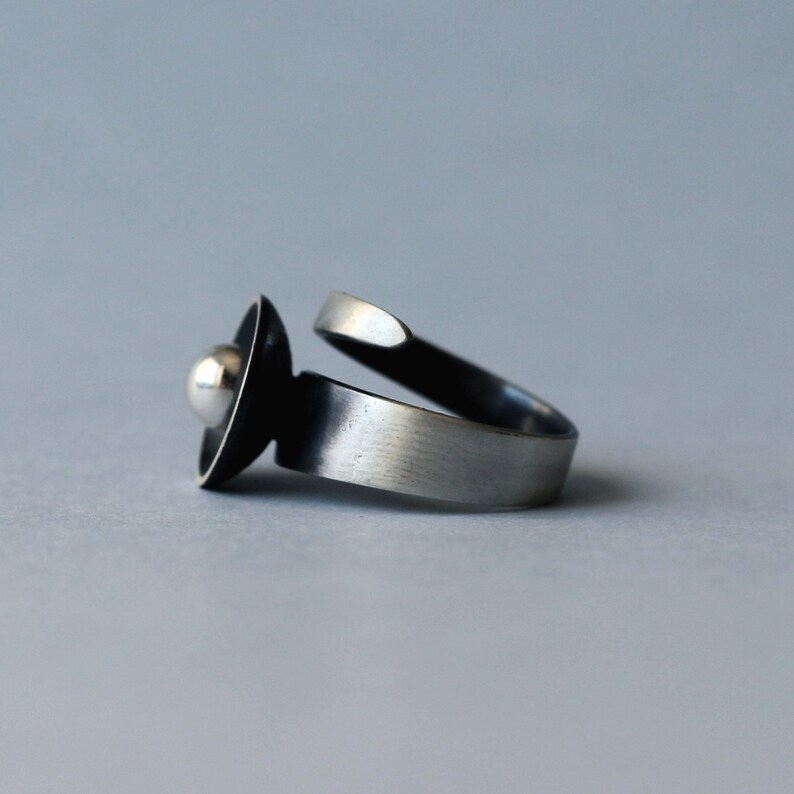 May include: A silver ring featuring a black and silver orb design. The ring is open and has a smooth, brushed finish.