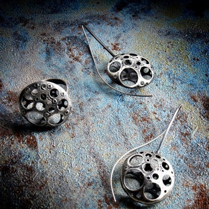 May include: Three silver earrings with a circular design featuring a pattern of small circles. The earrings are on a dark blue and brown textured background.