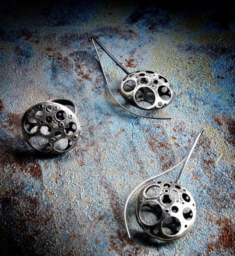 May include: Three silver earrings with a circular design featuring a pattern of small circles. The earrings are on a dark blue and brown textured background.