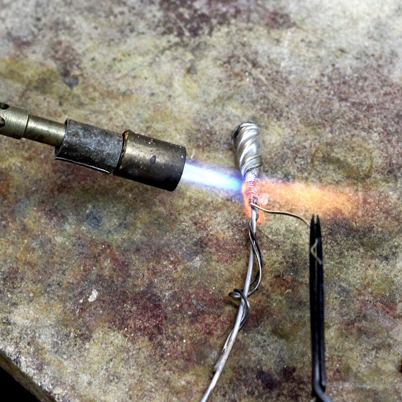 K&ouml;nnte beinhalten: Ein silbernes Metallobjekt wird mit einem L&ouml;tbrenner erhitzt. Das Objekt ist an einem Draht befestigt und wird mit einer Zange festgehalten. Die Flamme des L&ouml;tbrenners ist blau und orange.