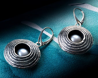 May include: A pair of silver earrings with a circular design and a pearl in the center. The earrings have a textured surface and a hook closure.