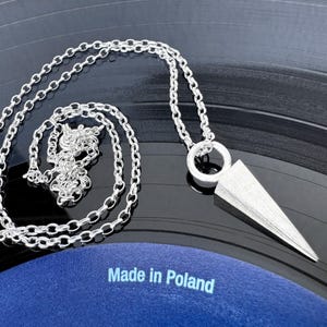 May include: A silver necklace featuring a geometric pendant. The pendant is a triangular shape with a textured finish. The chain is a delicate link style. The text "Made in Poland" is visible on a blue surface.