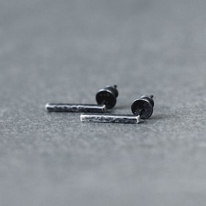Puede incluir: Un par de pendientes de barra de plata oxidada negra. Los pendientes están hechos de metal martillado y tienen un diseño simple y minimalista.