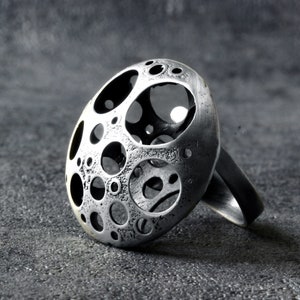 May include: A silver ring with a large, round, openwork design. The ring is made of metal and has a textured surface. The design features a series of circles and ovals, creating a unique and modern look.