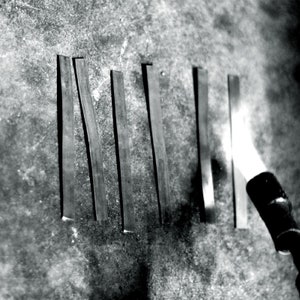 May include: A close-up of six metal strips being heated with a blowtorch. The metal strips are arranged vertically and are being heated from the bottom up. The blowtorch is in the lower right corner of the image.