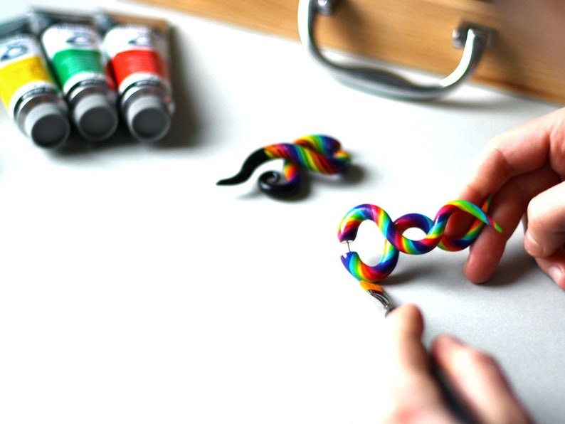 May include: Two rainbow colored spiral ear gauges. The gauges are made of polymer clay and are about 2 inches long. The gauges are on a white surface.