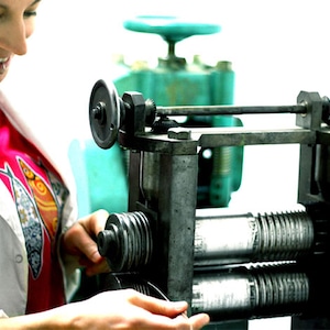 May include: A person operating a metal rolling mill machine. The machine has two large rollers and a smaller roller on the side. The person is feeding a thin metal strip into the machine.