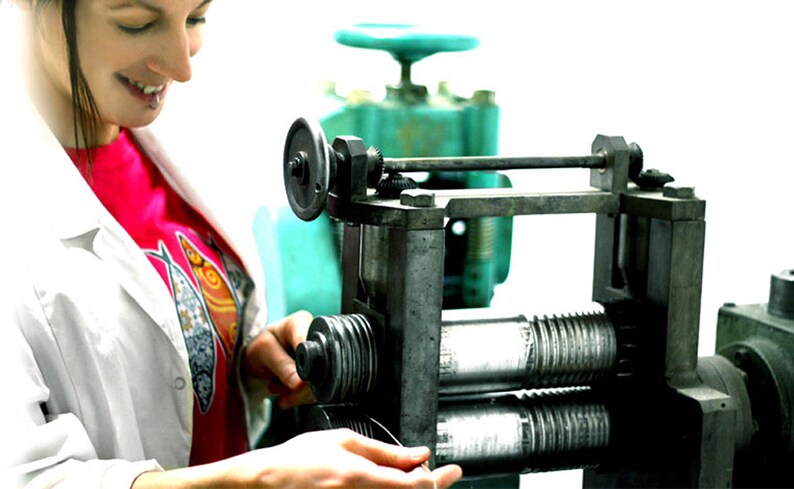 May include: A person operating a metal rolling mill machine. The machine has two large rollers and a smaller roller on the side. The person is feeding a thin metal strip into the machine.