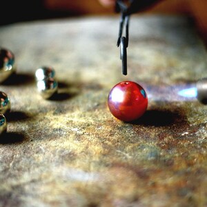 May include: A red glass bead is being heated with a blowtorch. The bead is being held in place by a pair of tweezers. There are several silver beads on the surface.