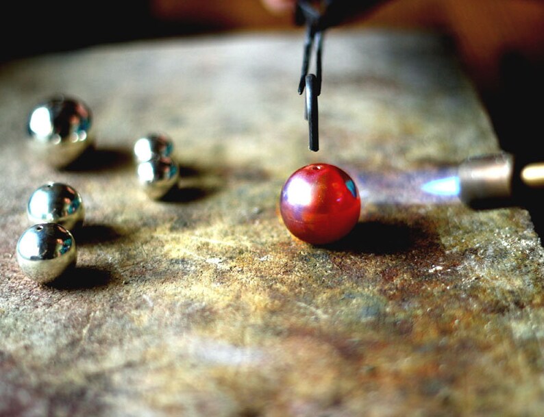 May include: A red glass bead is being heated with a blowtorch. The bead is being held in place by a pair of tweezers. There are several silver beads on the surface.